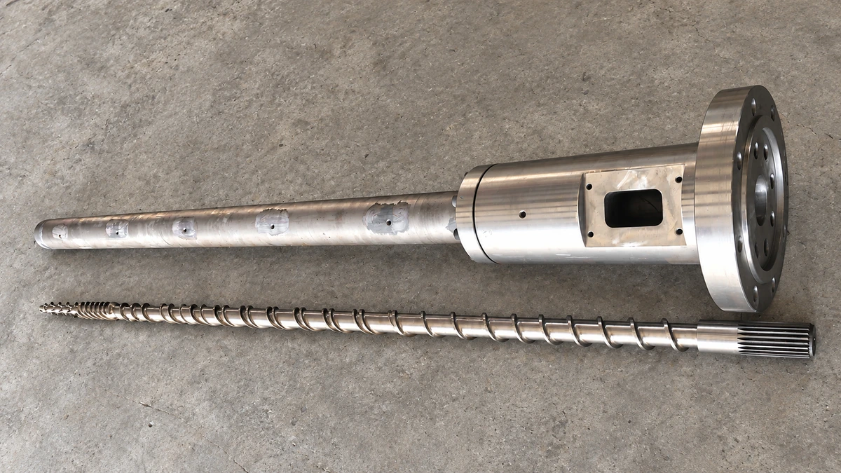 Single extruder screw beside a long flanged barrel with a rectangular feed opening on a gray floor